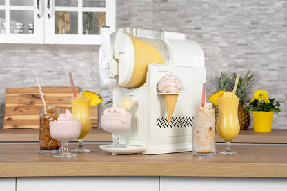 Die Unold Slusheismaschine Sofia, eine weiße Maschine für gefrorene Getränke auf einer modernen Küchentheke, gibt Softeis in eine Tüte aus. Um sie herum stehen Milchshakes und Eisgetränke mit Strohhalmen und Garnierungen.