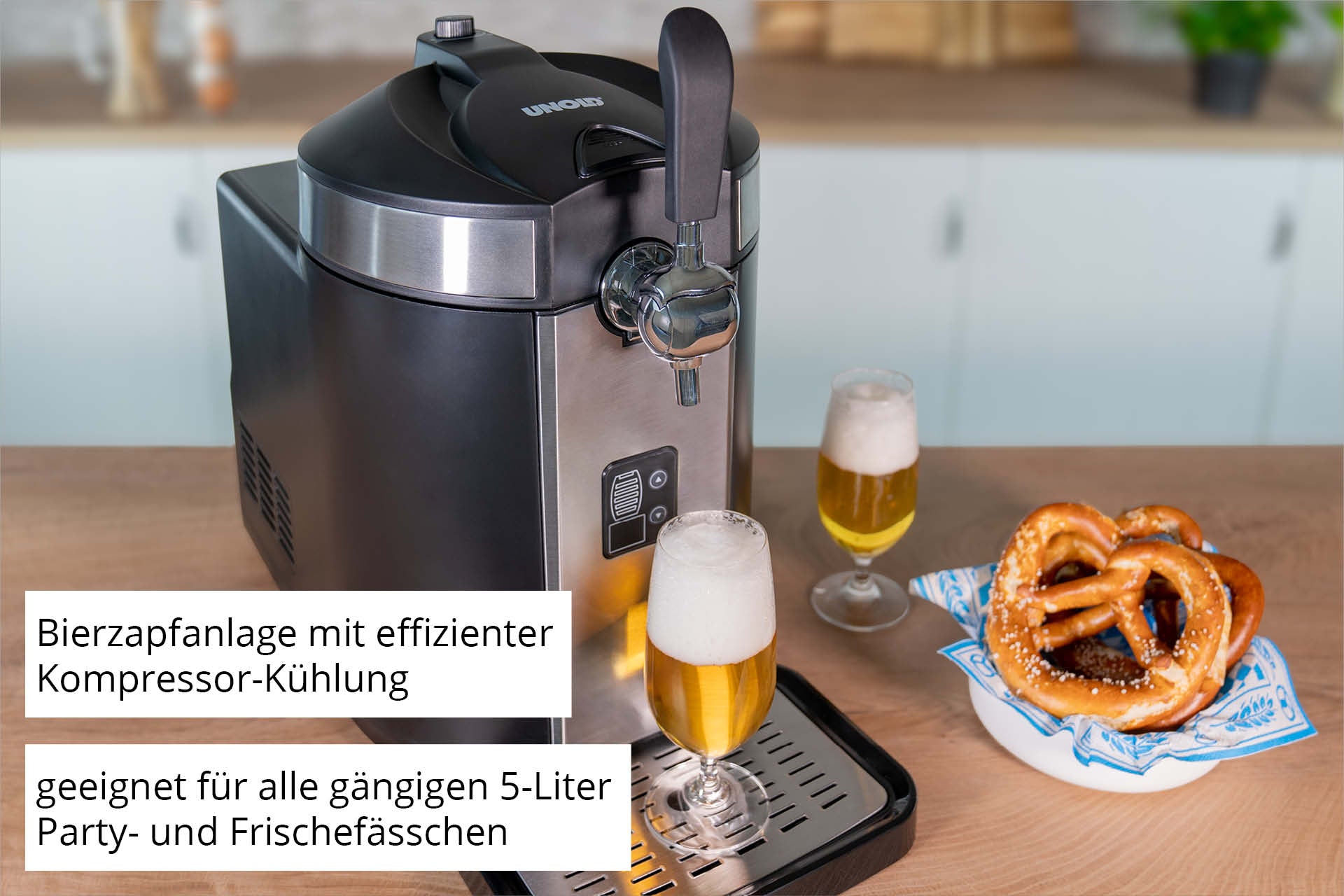 Die Unold Bierzapfanlage Franzl steht mit zwei gefüllten Biergläsern und einem Teller Brezeln auf der Küchentheke. Der deutsche Text hebt die effiziente Kompressorkühlung und die Kompatibilität mit 5-Liter-Fässern hervor.