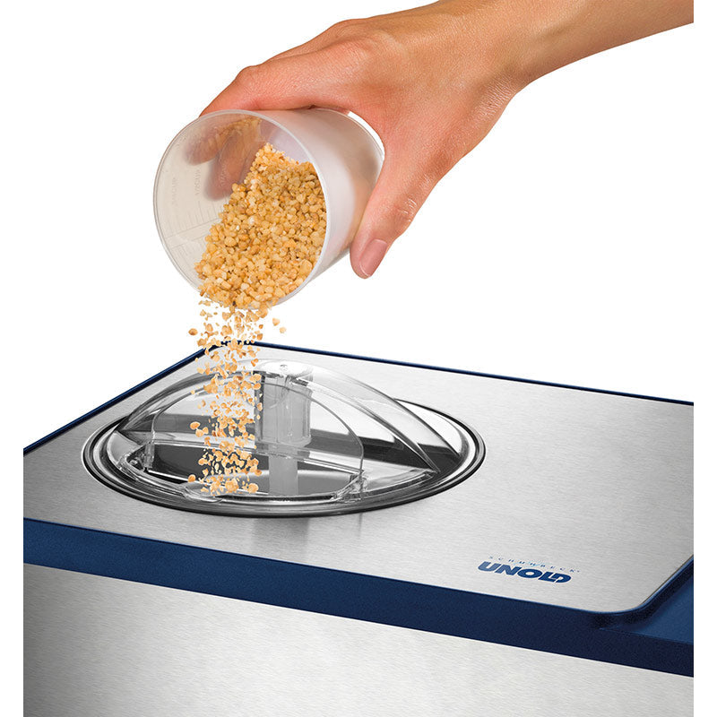 Eine Hand schüttet gehackte Nüsse aus einem kleinen Becher in die obere Öffnung der Unold Eismaschine Schuhbeck Exklusiv inklusive Eisbuch 1,5 L, einer Eismaschine aus Edelstahl mit transparentem Deckel.