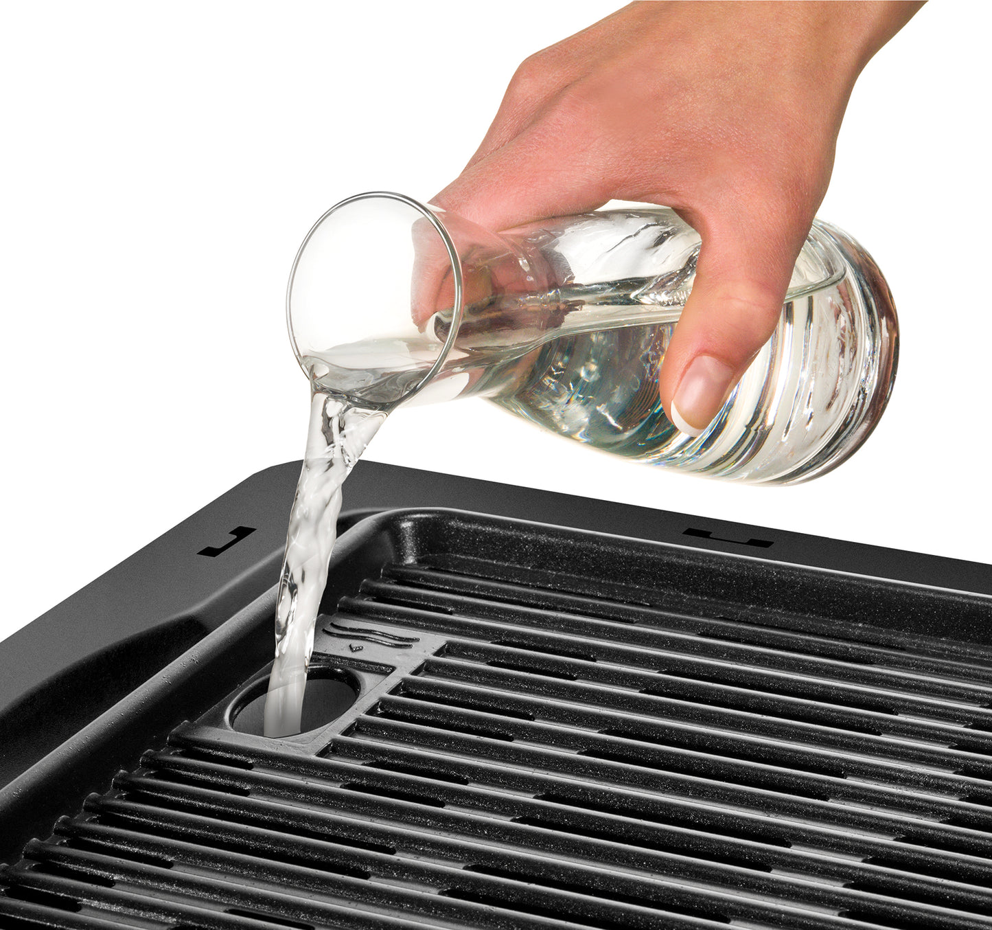 Eine Hand gießt Wasser aus einem Glaskrug in die dafür vorgesehene Öffnung des Unold 58565 Barbecue-Grill Vario, im Vordergrund sind Grillroste des schwarzen Unold Indoor-Grills zu sehen.