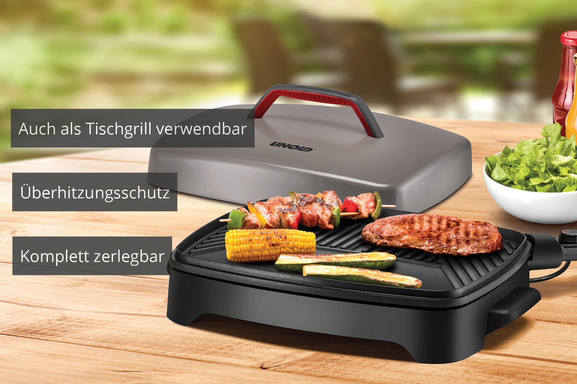 Der Unold Barbecue Power Grill steht auf einer Holzplatte und brutzelt mit Spießen, Mais und Steak. Der Deckel ist seitlich angebracht. Der deutsche Text weist auf die Tischnutzung, den Überhitzungsschutz und die einfache Demontage hin.