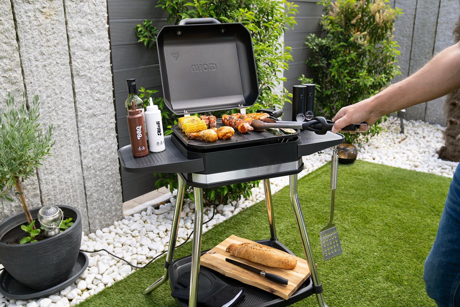 Eine Person grillt Mais, Würstchen und Hühnchen auf einem schwarzen Grill in einem Garten. Ein Laib Brot, Soßen und Grillbesteck liegen in der Nähe. Grüne Pflanzen und weiße Steine umgeben den Bereich.