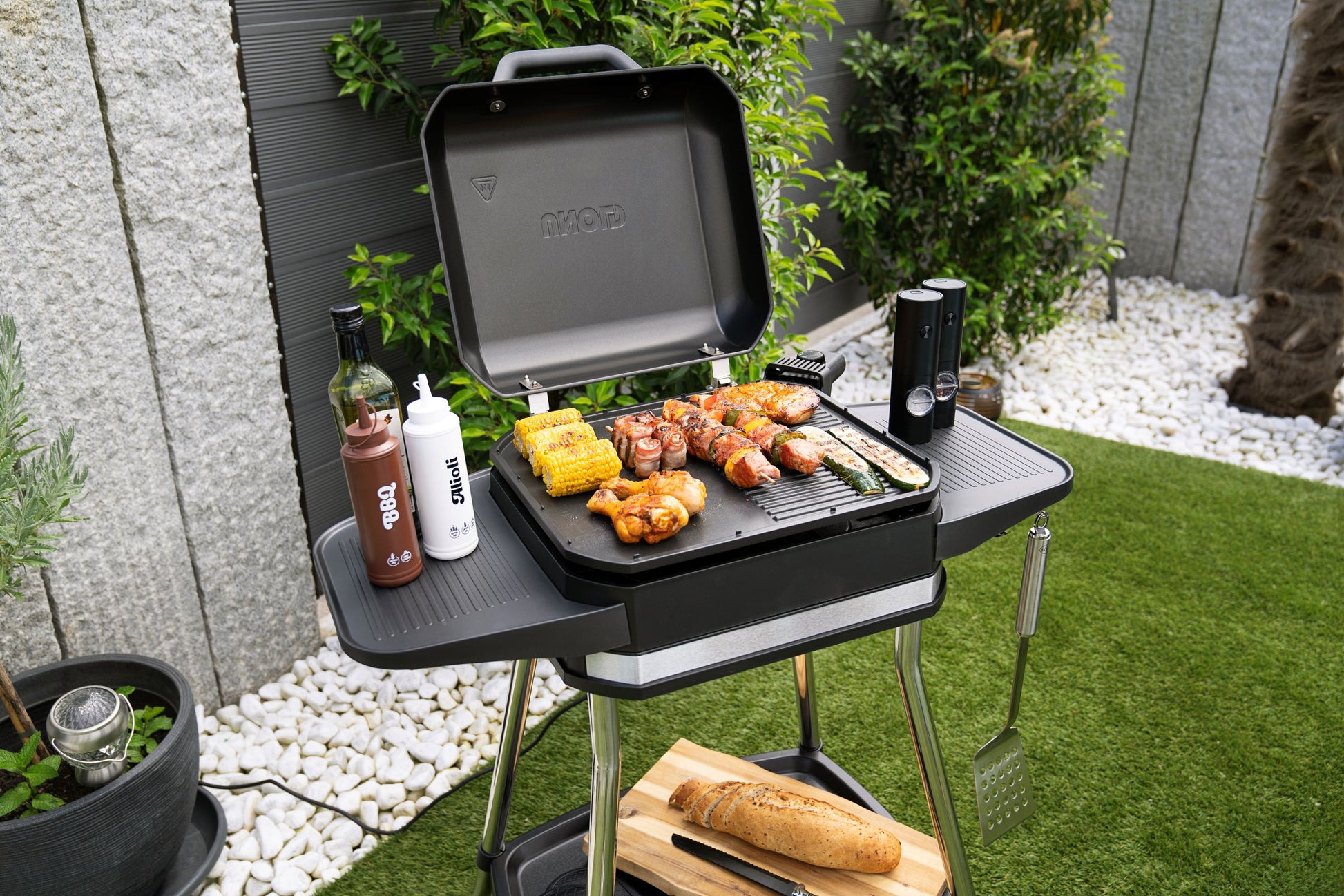 Der Unold BBQ-Stand-/Tischgrill Jimmy grillt Fleisch, Mais und Gemüse auf einer Terrasse. BBQ-Saucen, eine Pfeffermühle, Brot auf einem Schneidebrett und ein Pfannenwender liegen bereit. Grüne Pflanzen und weiße Steine runden die Szene ab.