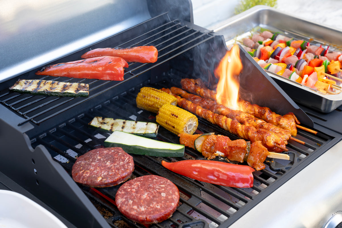 Der Unold Barbecue Grill-Wagen Buddy wird gezeigt, wie er Burger, Mais, Zucchini, rote Paprika, Lachs und Hähnchenspieße mit einer kleinen Flamme darunter grillt; daneben steht ein Tablett mit bunten Gemüse- und Fleischspießen.