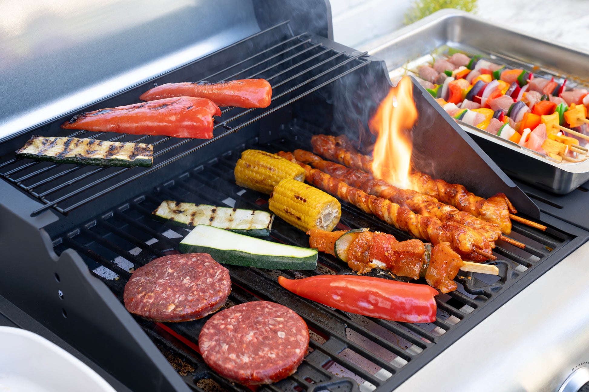 Der Unold Barbecue Grill-Wagen Buddy wird gezeigt, wie er Burger, Mais, Zucchini, rote Paprika, Lachs und Hähnchenspieße mit einer kleinen Flamme darunter grillt; daneben steht ein Tablett mit bunten Gemüse- und Fleischspießen.