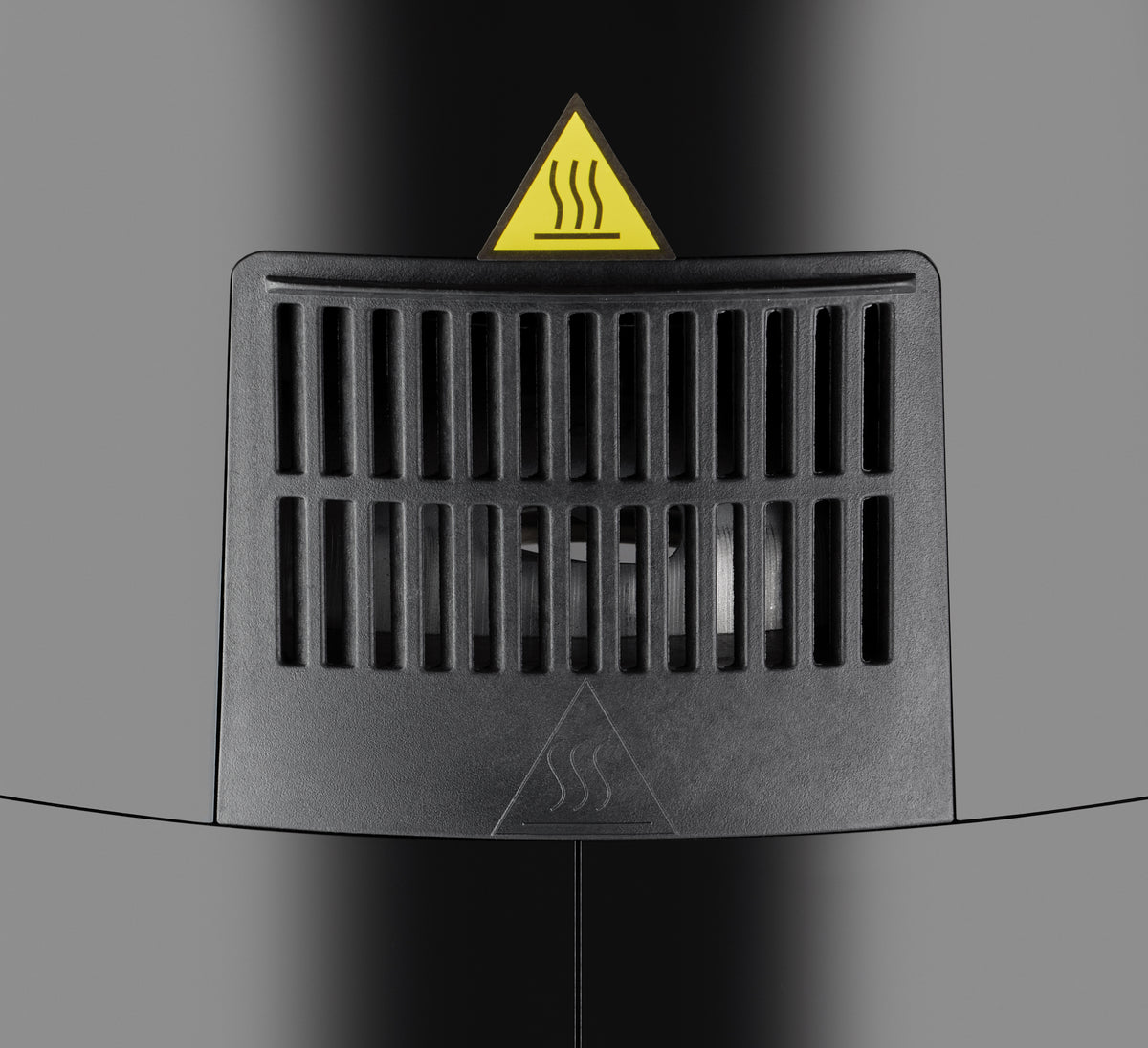Nahaufnahme einer schwarzen Entlüftungsöffnung mit vertikalen Lamellen an der Unold Heißluftfritteuse XL. Darüber befindet sich ein dreieckiges Warnschild mit Hitzewellensymbol, das auf heiße Luft oder eine Wärmequelle hinter der Entlüftung hinweist.