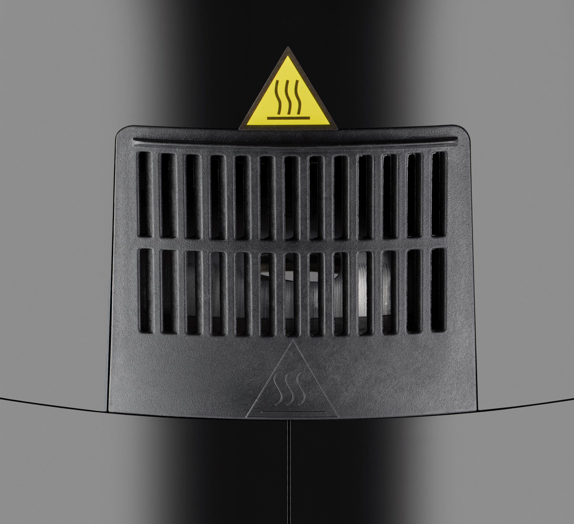 Nahaufnahme einer schwarzen Entlüftungsöffnung mit vertikalen Lamellen an der Unold Heißluftfritteuse XL. Darüber befindet sich ein dreieckiges Warnschild mit Hitzewellensymbol, das auf heiße Luft oder eine Wärmequelle hinter der Entlüftung hinweist.