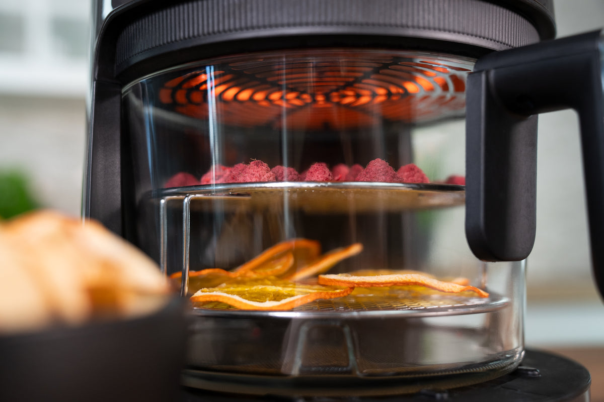 Nahaufnahme des Heißluftfritteuse-Dörrgeräts „Unold Heißluftfritteuse Glas“ mit Glastür, auf der Tabletts mit geschnittenen Orangen und Himbeeren zu sehen sind, die unter einem leuchtend orangefarbenen Heizelement trocknen.