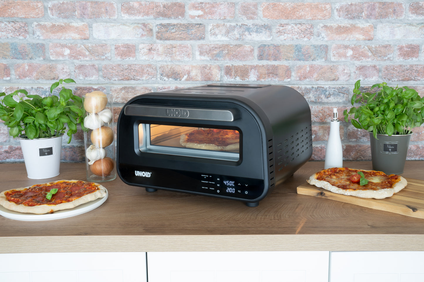Ein schwarzer Unold Pizzaofen Don Luigi steht auf einer Holztheke und backt eine Pizza. Um ihn herum stehen frische Pizzen, Basilikumpflanzen, Zwiebeln, Salz und Pfeffer vor einer Backsteinmauer.