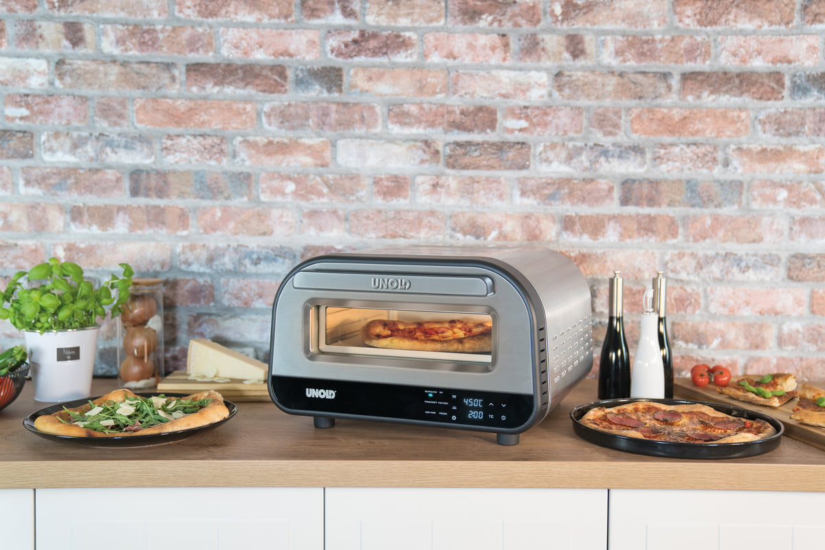 Der Unold Pizzaofen LUIGI steht kompakt auf einer Küchentheke mit einer Backsteinwand im Hintergrund. Frisch gebackene Pizzen liegen neben dem Ofen, umgeben von Kräutern, Gewürzen und Gemüse für eine schmackhafte Note.