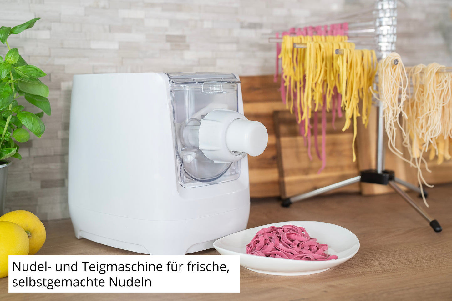 Die Unold Nudelmaschine Nora, eine weiße Nudel- und Teigmaschine, steht auf einer Küchentheke neben violetten Nudeln, gelben Nudeln auf einem Trockengestell, Zitronen und einem Holzbrett. Ein deutscher Text beschreibt die Maschine.