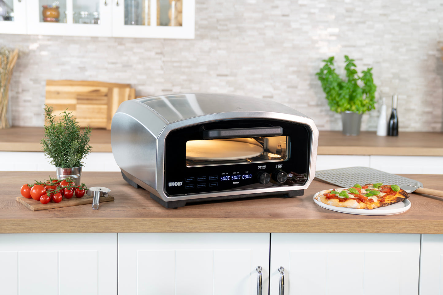 Der Unold Pizzaofen Enzo sitzt auf einer modernen Küchenzeile und backt eine Pizza. Frische Kräuter, Kirschtomaten, ein Pizzaschneider und eine fertige Pizza auf einer Schiene liegen in der Nähe, alles in einer sauberen, modernen Küche.