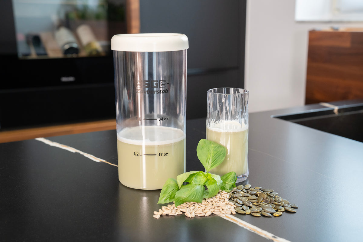 Der ESGE-Zauberstab Magic Fusion Mixer von ESGE Zauberstab steht mit seinem mit einer hellgrünen Flüssigkeit gefüllten Glasbehälter auf einer Küchentheke, neben frischen Basilikumblättern, Sonnenblumenkernen und Kürbiskernen.