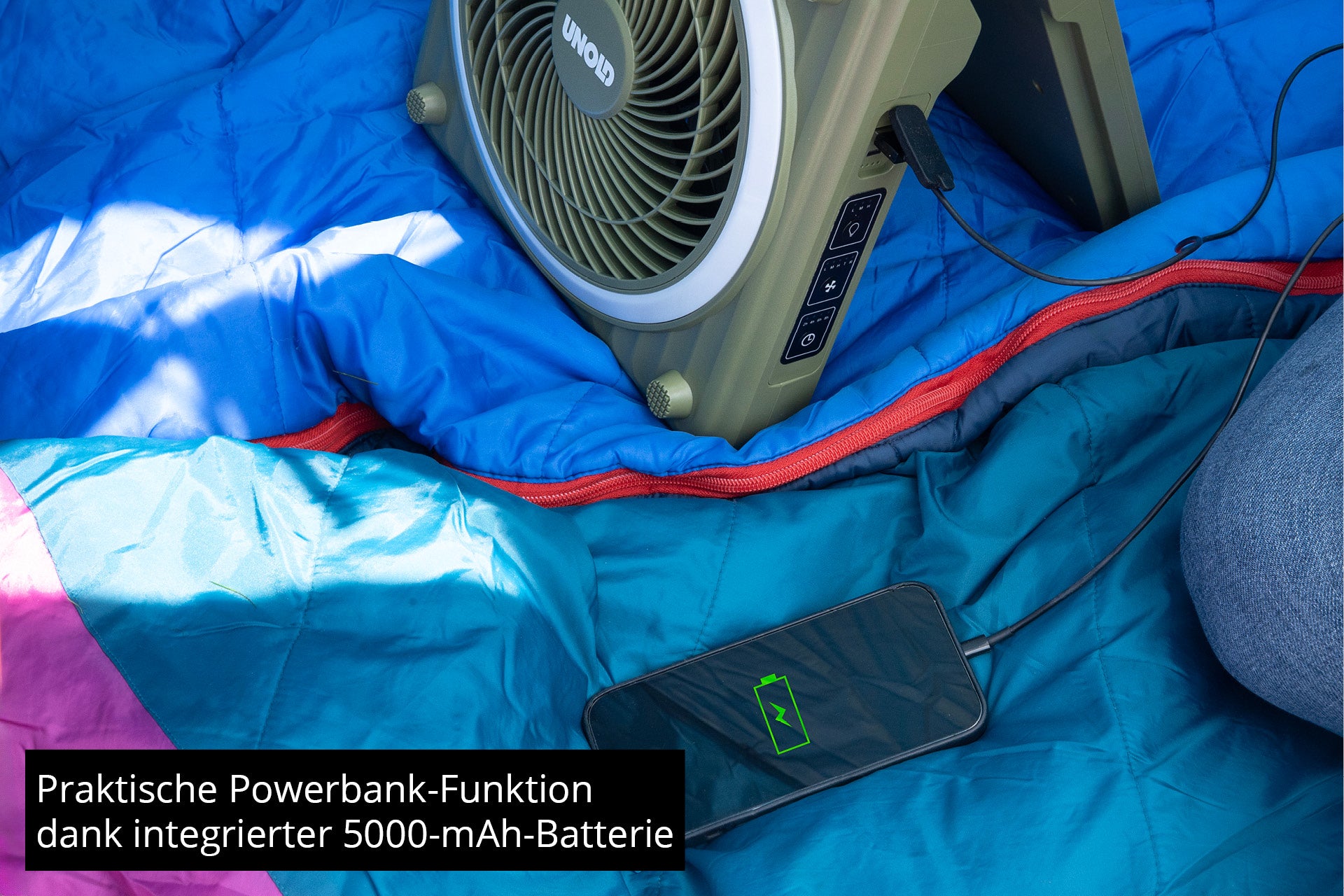 Ein Solarventilator Camping von Unold lädt ein Smartphone per Kabel an den USB-Ports auf, während er auf einem blauen Schlafsack liegt. Deutschsprachiger Text hebt die Powerbank-Funktion und den integrierten 5000-mAh-Akku hervor.