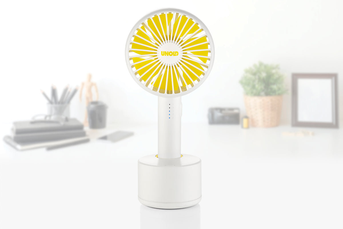 Ein kompaktes weiß-gelbes Unold Ventilatoren-Set Tischventilator Breezy Clip + Handventilator Breezy Swing steht auf einem Schreibtisch, mit verschwommenen Büromaterialien, einer kleinen Pflanze und einem Bilderrahmen im Hintergrund.