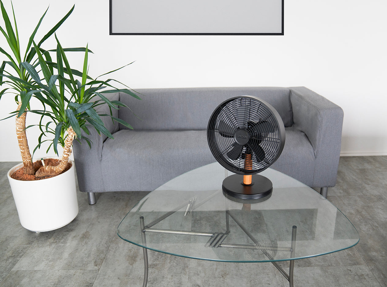 Der Unold Tischventilator Black Steel steht auf einem Couchtisch aus Glas vor einem grauen Sofa, links davon steht eine große Topfpflanze, alles in einem modernen, minimalistischen Raum mit grauem Boden.