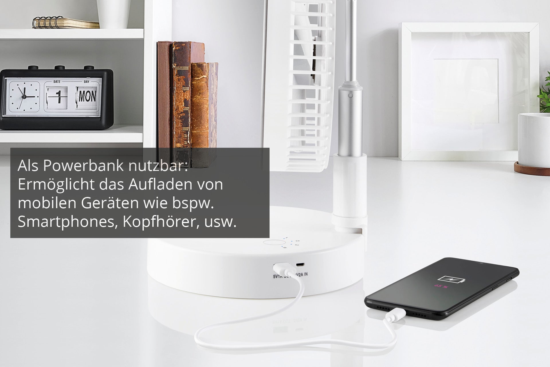 Ein Smartphone lädt per Kabel an der Basisstation des weißen Unold TELESKOP-VENTI CORDLESS Standventilators. Im Hintergrund sind Uhr, Kalender und Bücher auf einem weißen Regal. Ein Textfeld erklärt die Powerbank-Funktion der Basis.