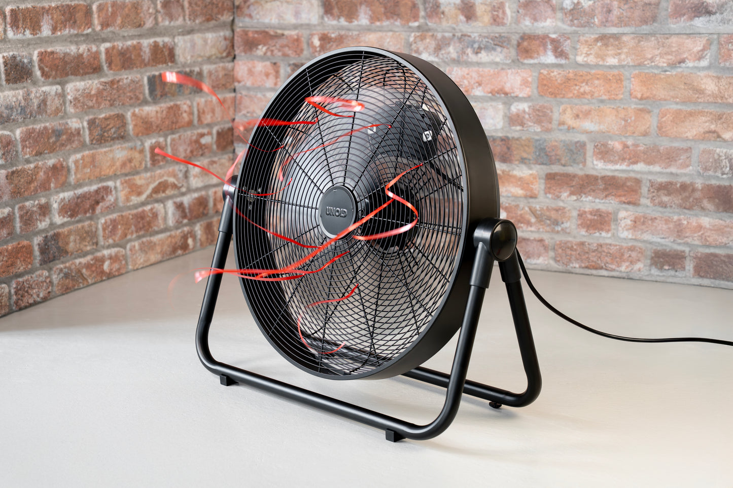 Der elektrische Bodenventilator Unold Windmaschine Black Steel, der nach oben gerichtet ist, bläst rote Bänder, um den Luftstrom in einem Raum mit hellgrauem Fußboden und Sichtziegelwänden zu zeigen. Der schwarze Ventilator ist eingesteckt.
