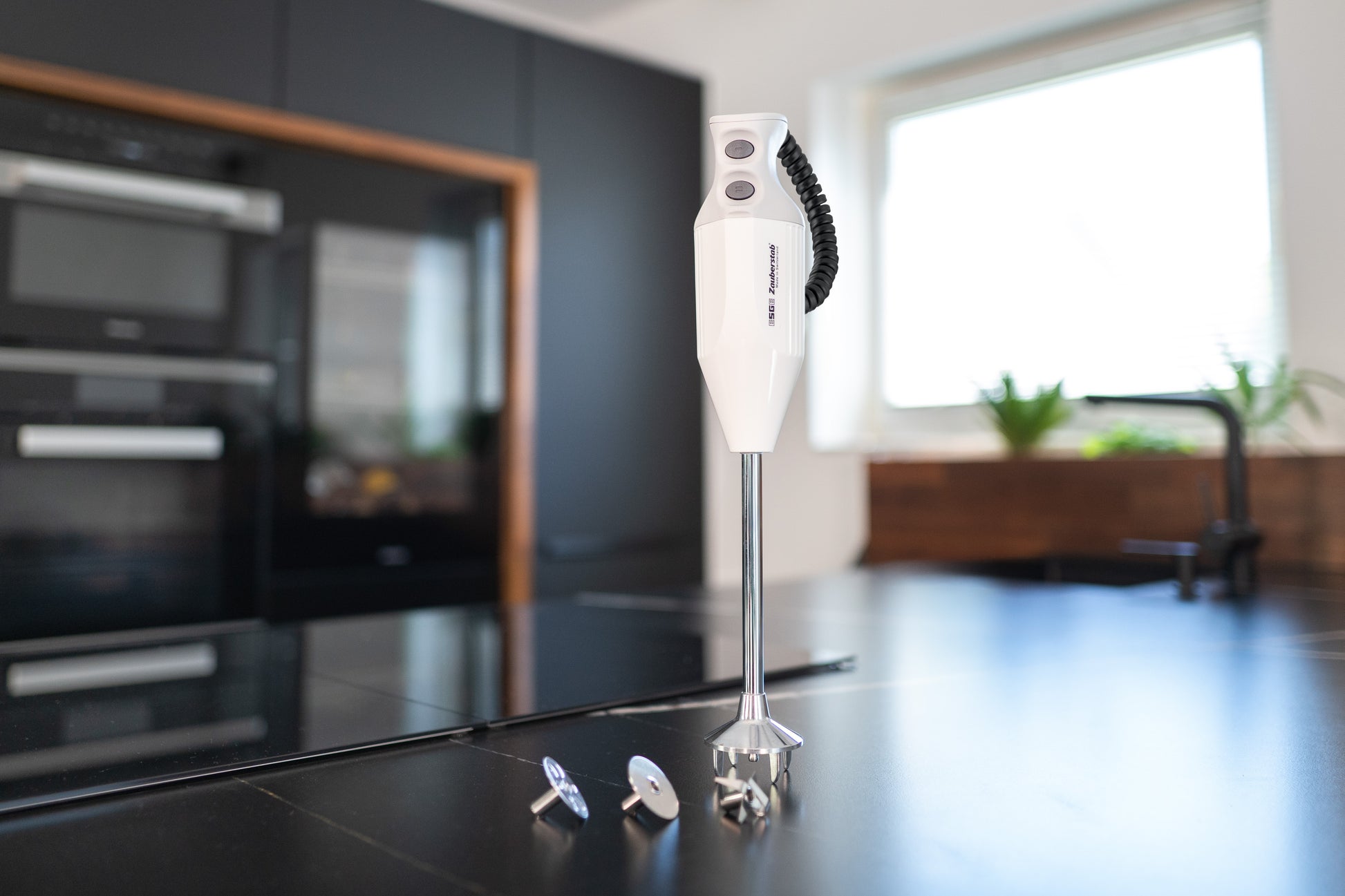 Der Unold ESGE-Zauberstab® P 200 Profi, ein weißer Stabmixer, steht aufrecht auf einem schwarzen Küchentisch mit seinen Metallaufsätzen in der Nähe in einer modernen Küche mit schwarzen Schränken und Einbaugeräten.