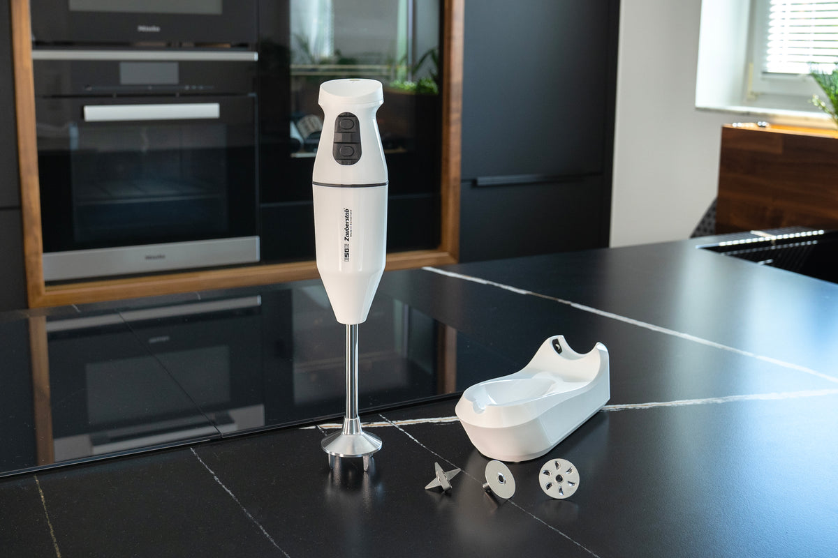 Der kabellose Stabmixer Unold ESGE-Zauberstab® Weiß 95100 mit Messer- und Schneebesenaufsatz steht aufrecht auf einer schwarzen Küchenarbeitsplatte, begleitet von einer weißen Ablagefläche und modernen Geräten im Hintergrund.