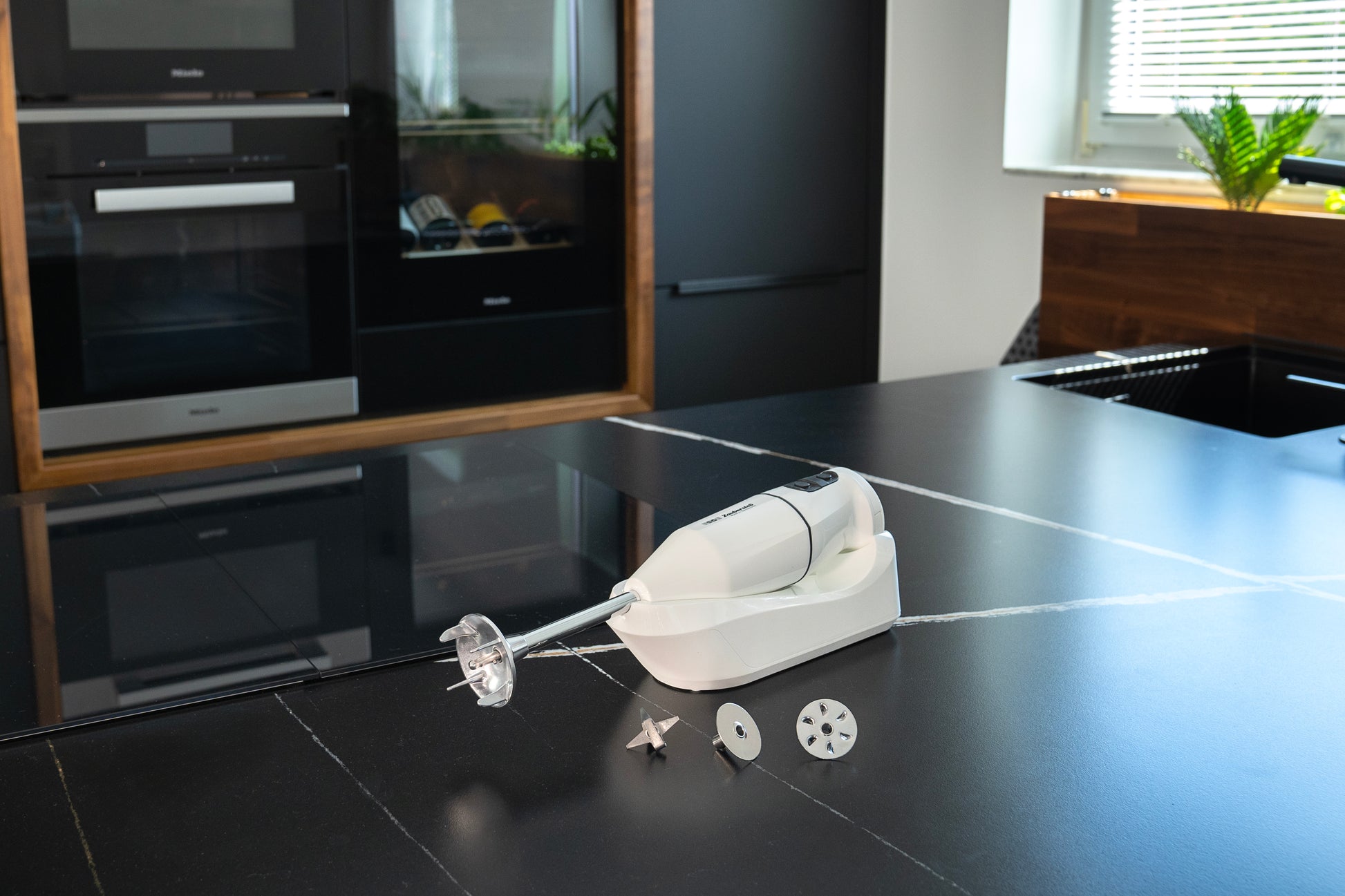 Der kabellose Handmixer Unold ESGE-Zauberstab® Weiß 95100 mit Aufsätzen steht auf einer schwarzen Küchenarbeitsplatte, im Hintergrund sind moderne Geräte und eine Pflanze zu sehen.