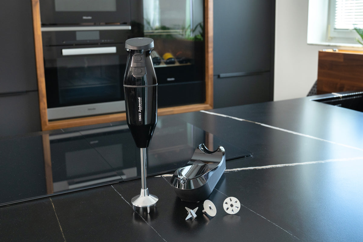 Der kabellose Stabmixer Unold ESGE-Zauberstab® Schwarz 95105 mit Aufsätzen und Ständer wird auf einer modernen schwarzen Küchentheke gezeigt, im Hintergrund sind Einbaubacköfen und ein Fenster zu sehen.