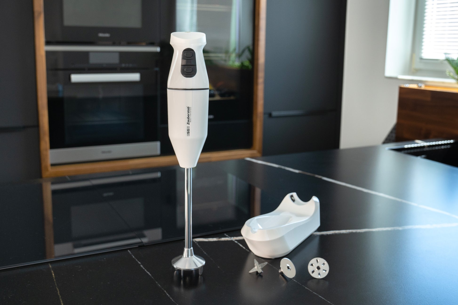 Der kabellose Stabmixer Unold ESGE-Zauberstab® PRO Weiß 95300 mit Standfuß und zwei Metallaufsätzen steht auf einer eleganten schwarzen Küchentheke. Im Hintergrund sind Einbauherde, Schränke und ein sonnenbeschienenes Fenster zu sehen.
