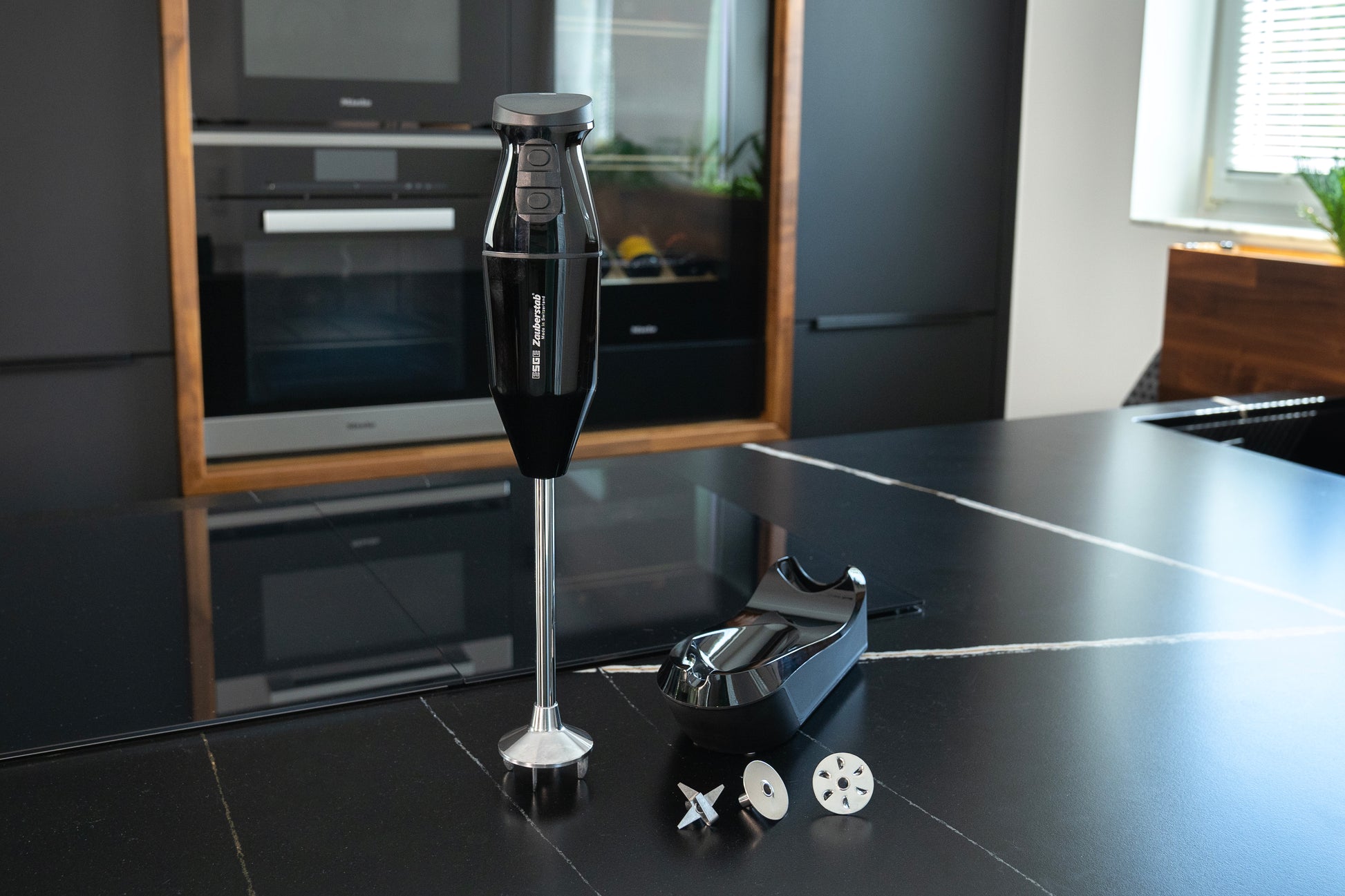 Der kabellose Stabmixer Unold ESGE-Zauberstab® PRO Schwarz 95305 mit Standfuß und drei Messeraufsätzen steht auf einer schlichten, schwarzen Küchenarbeitsplatte. Im Hintergrund sind Einbaubacköfen und ein Fenster zu sehen.