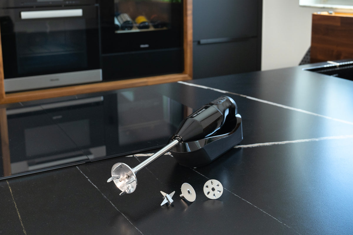 Der kabellose Stabmixer Unold ESGE-Zauberstab® PRO Schwarz 95305 mit Metallklingenaufsätzen und Aufbewahrungskoffer steht auf einer eleganten schwarzen Küchentheke in einer modernen Küche.