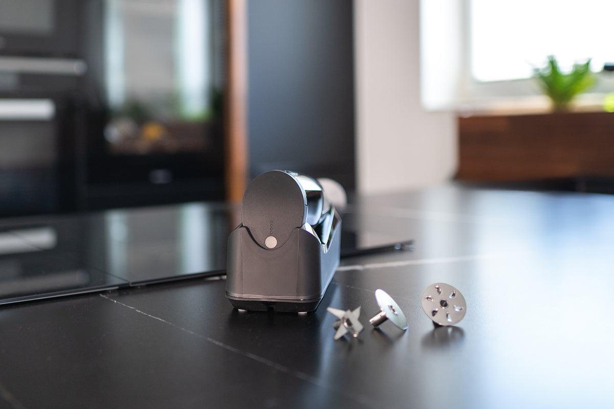 Eine Nahaufnahme des Unold ESGE-Zauberstab® Akku PRO Schwarz 95305 auf einer schwarzen Arbeitsplatte, präsentiert mit mehreren Metallaufsätzen. Im Hintergrund ist eine moderne Küche mit verschwommenen Geräten und einem Fenster zu sehen.