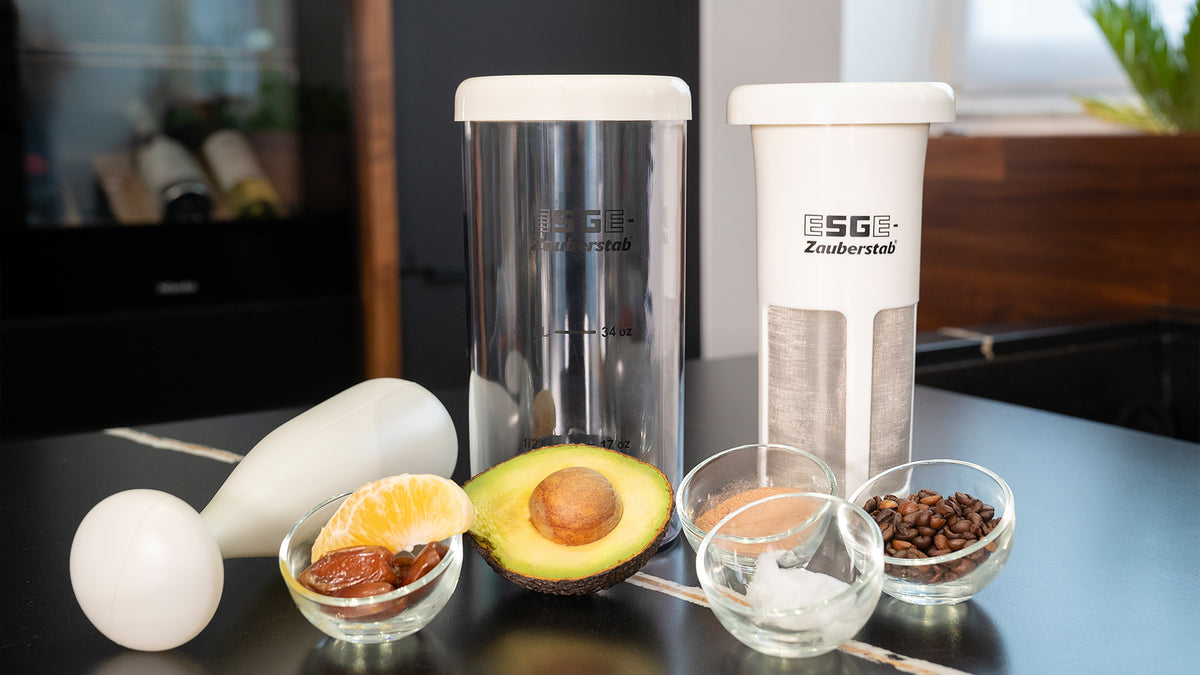 Auf einer Küchentheke stehen zwei ESGE Zauberstab-Mixer mit Behältern, umgeben von kleinen Schüsseln mit Datteln, einer Orangenscheibe, Avocado, Kokosöl und Kaffeebohnen.