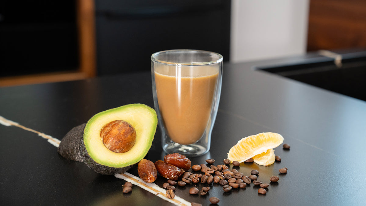Auf einer schwarzen Theke steht ein Glas brauner Smoothie, umgeben von einer halben Avocado, Kaffeebohnen, Datteln und einer Orangenspalte.