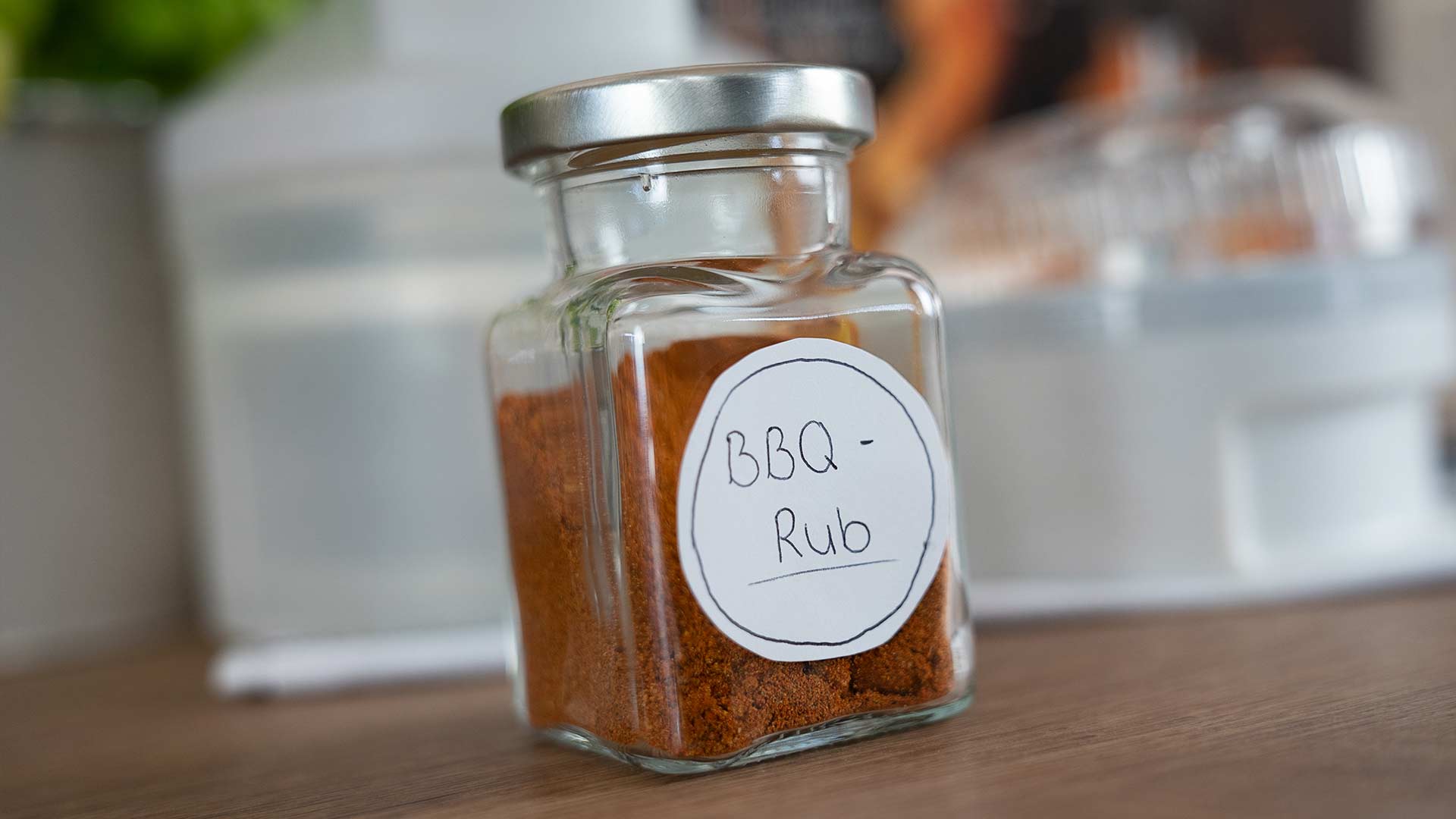 Ein Glas mit silbernem Deckel und der Aufschrift „BBQ Rub“ steht auf einer Holzfläche. Das Glas ist mit einer rotbraunen Gewürzmischung gefüllt. Im Hintergrund sind verschwommene Küchenutensilien zu sehen.