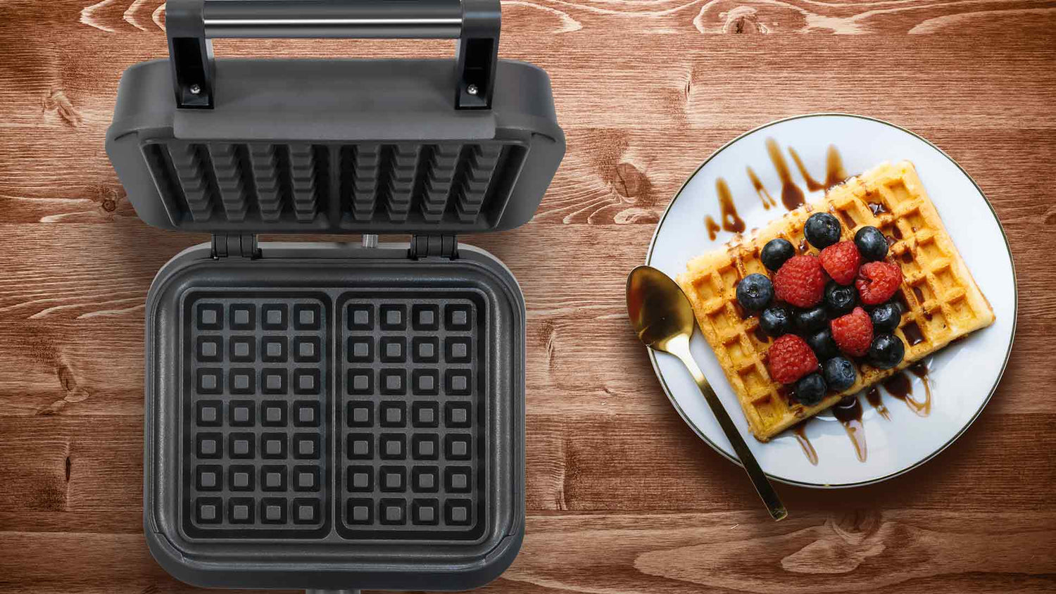 Auf einem Holztisch steht ein geöffnetes Waffeleisen neben einem Teller mit einer Waffel, die mit Blaubeeren, Himbeeren und Schokoladensoße garniert ist, und neben dem Teller liegt ein goldener Löffel.