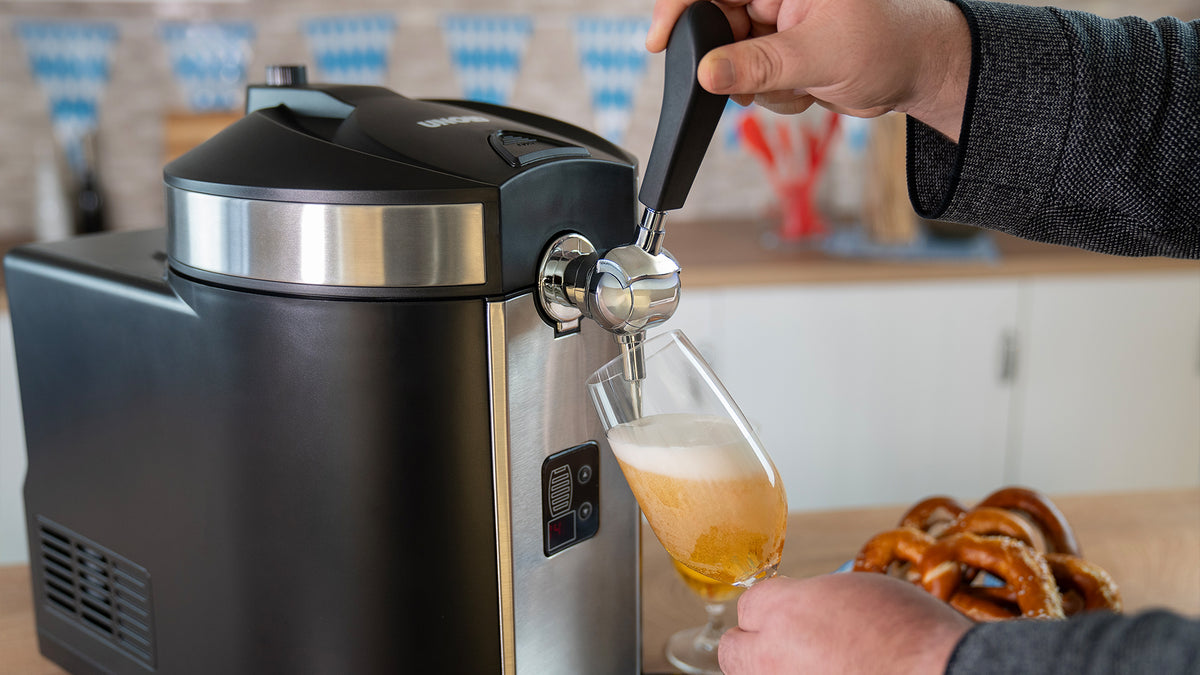 Eine Person schenkt in einer Küche Bier aus einem Zapfhahn in ein Glas ein. Daneben liegen weiche Brezeln auf einem Teller.