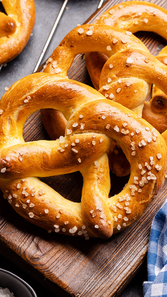 Eine Nahaufnahme von goldbraunen, weichen Brezeln, bestreut mit grobem Salz, die auf einem Holzbrett liegen, daneben liegt ein blau kariertes Tuch.