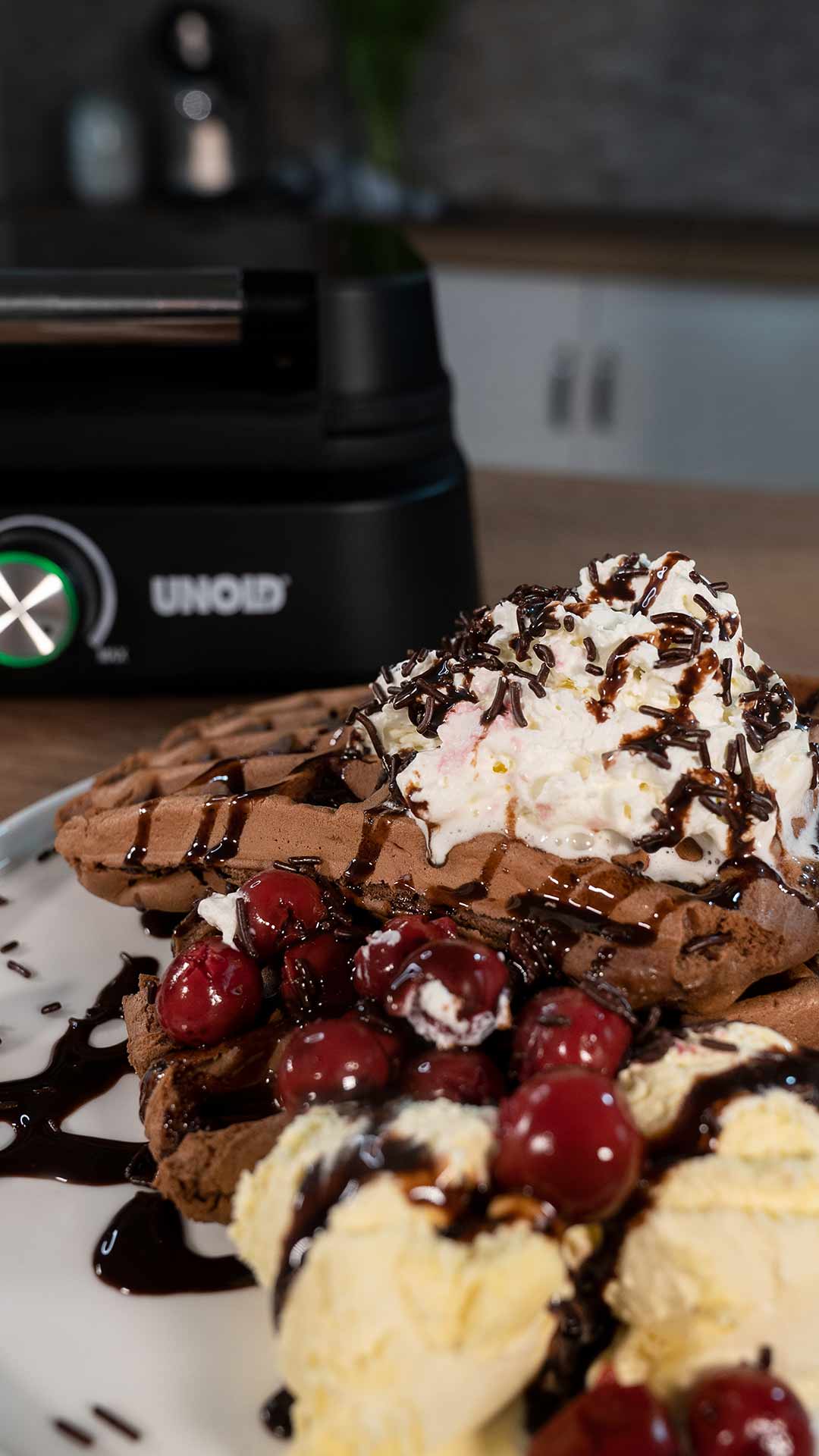 Ein Teller mit Schokoladenwaffeln, garniert mit Schlagsahne, Schokoladensirup, Kirschen und Vanilleeis, im Hintergrund ein Waffeleisen.