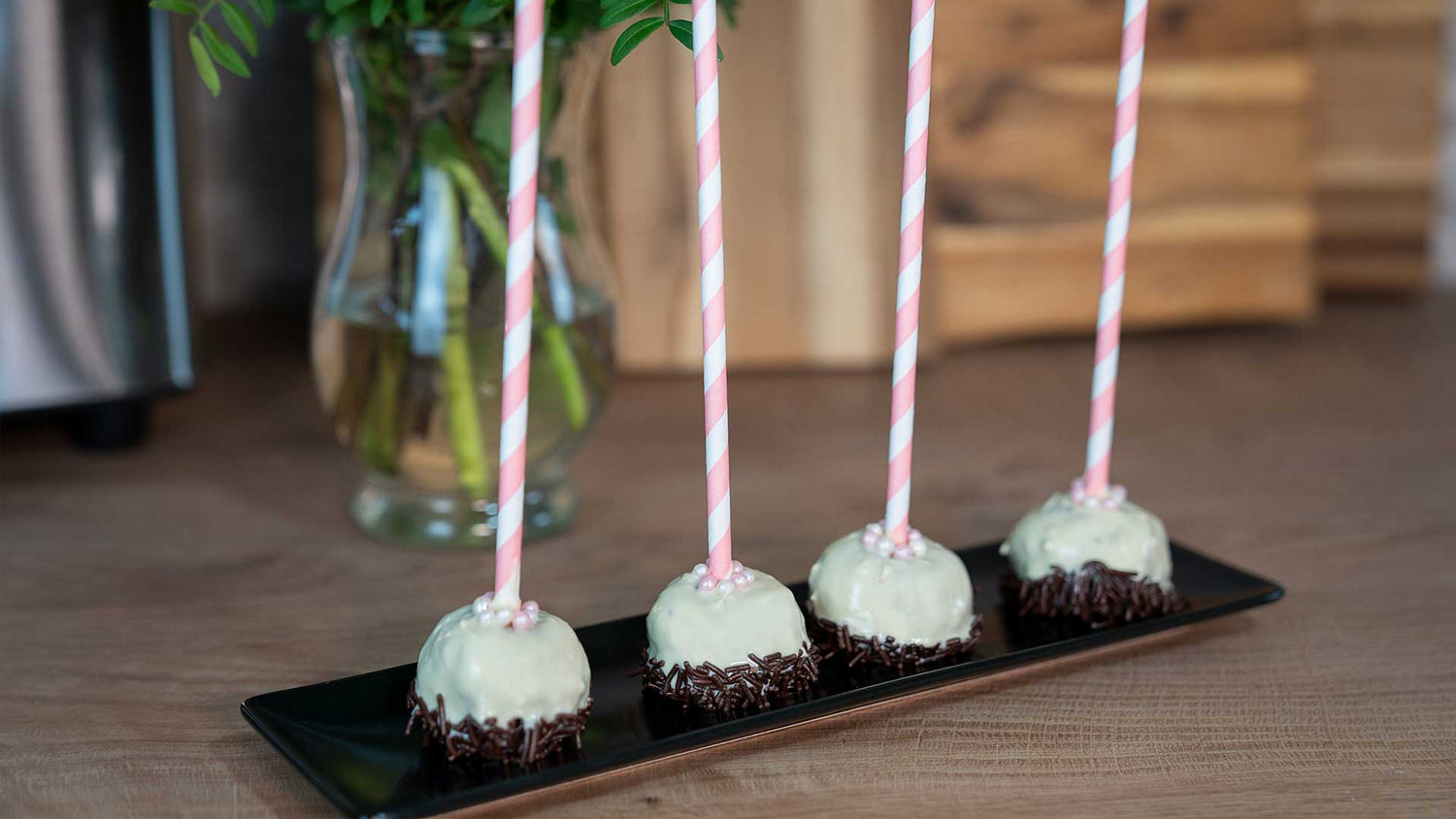 Vier Cake Pops mit rosa-weiß gestreiften Stielen, überzogen mit weißer Schokolade und Schokostreuseln, liegen in einer Reihe auf einem schwarzen rechteckigen Teller auf einer Holzoberfläche. Im Hintergrund steht eine Glasvase mit Blättern.