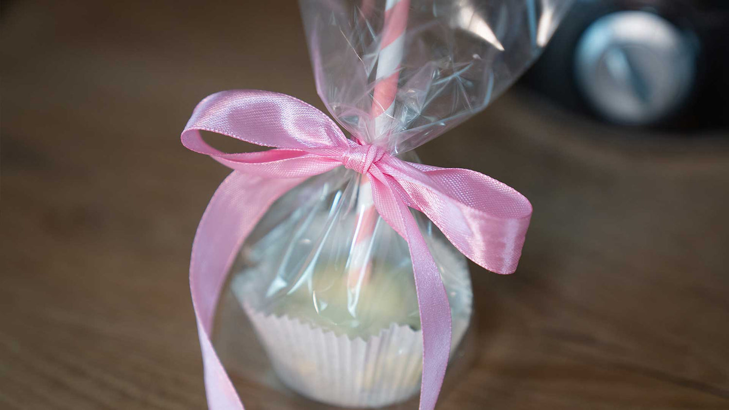 Ein in durchsichtige Plastikfolie eingewickelter Cupcake mit einer rosa Schleife, die um einen Stiel gebunden ist, liegt auf einer Holzoberfläche.