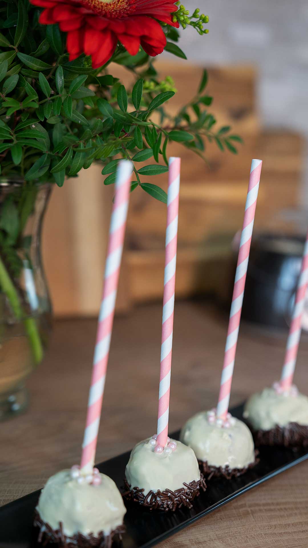 Vier Cake Pops mit weißem Schokoladenüberzug, Schokoladenstreuseln am Boden und rosa gestreiften Papierstrohhalmen stehen auf einem schwarzen Tablett, im Hintergrund steht eine Vase mit roten Blumen und Grünpflanzen.