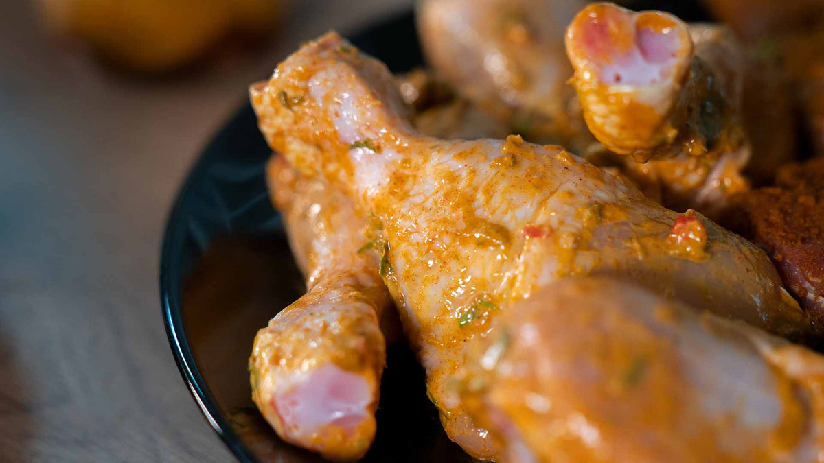 Nahaufnahme von rohen Hähnchenkeulen, überzogen mit einer gelblichen Marinade mit Kräutern und Gewürzen, auf einem schwarzen Teller.