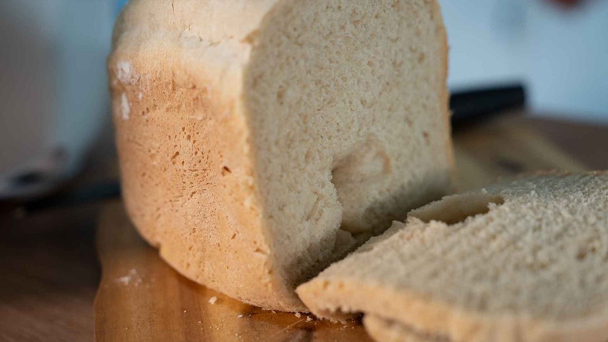 Ein Laib selbstgebackenes Brot mit goldbrauner Kruste liegt auf einem Holzschneidebrett. Es ist teilweise angeschnitten und zeigt ein weiches, luftiges Inneres. Im Hintergrund ist ein Brotmesser zu sehen.