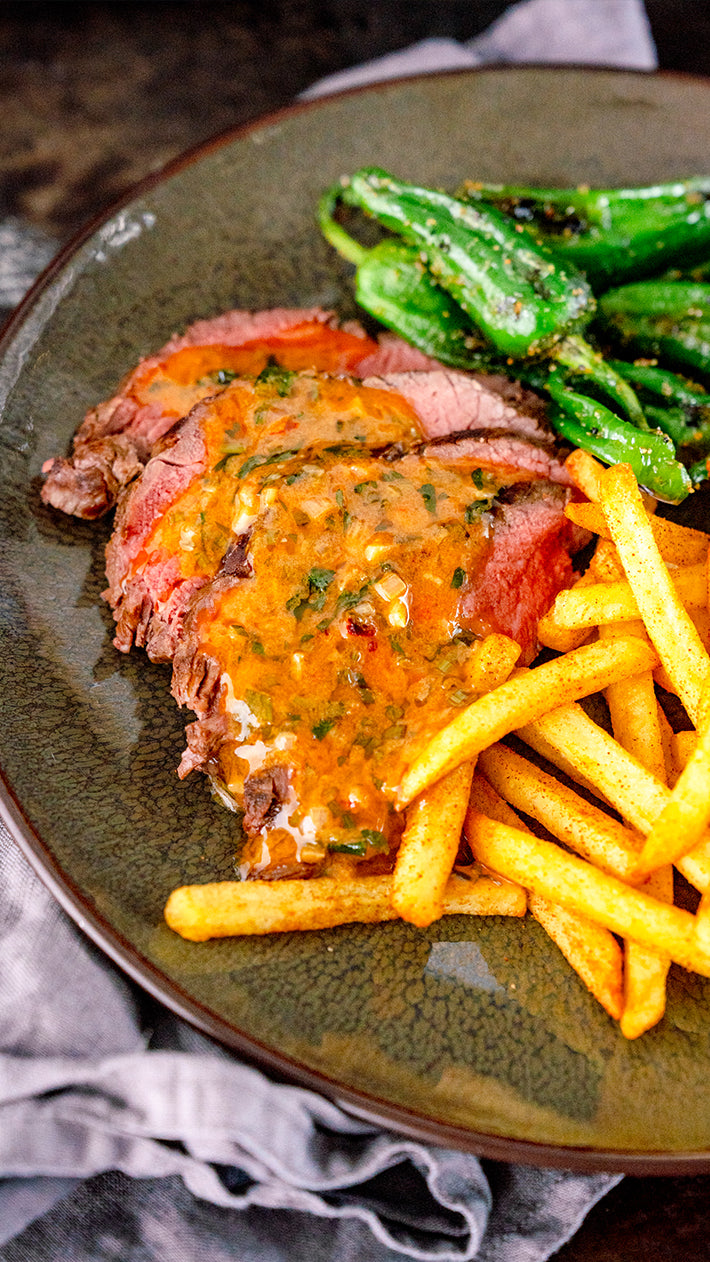 Ein Teller mit Steakscheiben, garniert mit einer cremigen Kräutersauce, serviert mit gewürzten Pommes Frites und sautierten grünen Paprikaschoten.