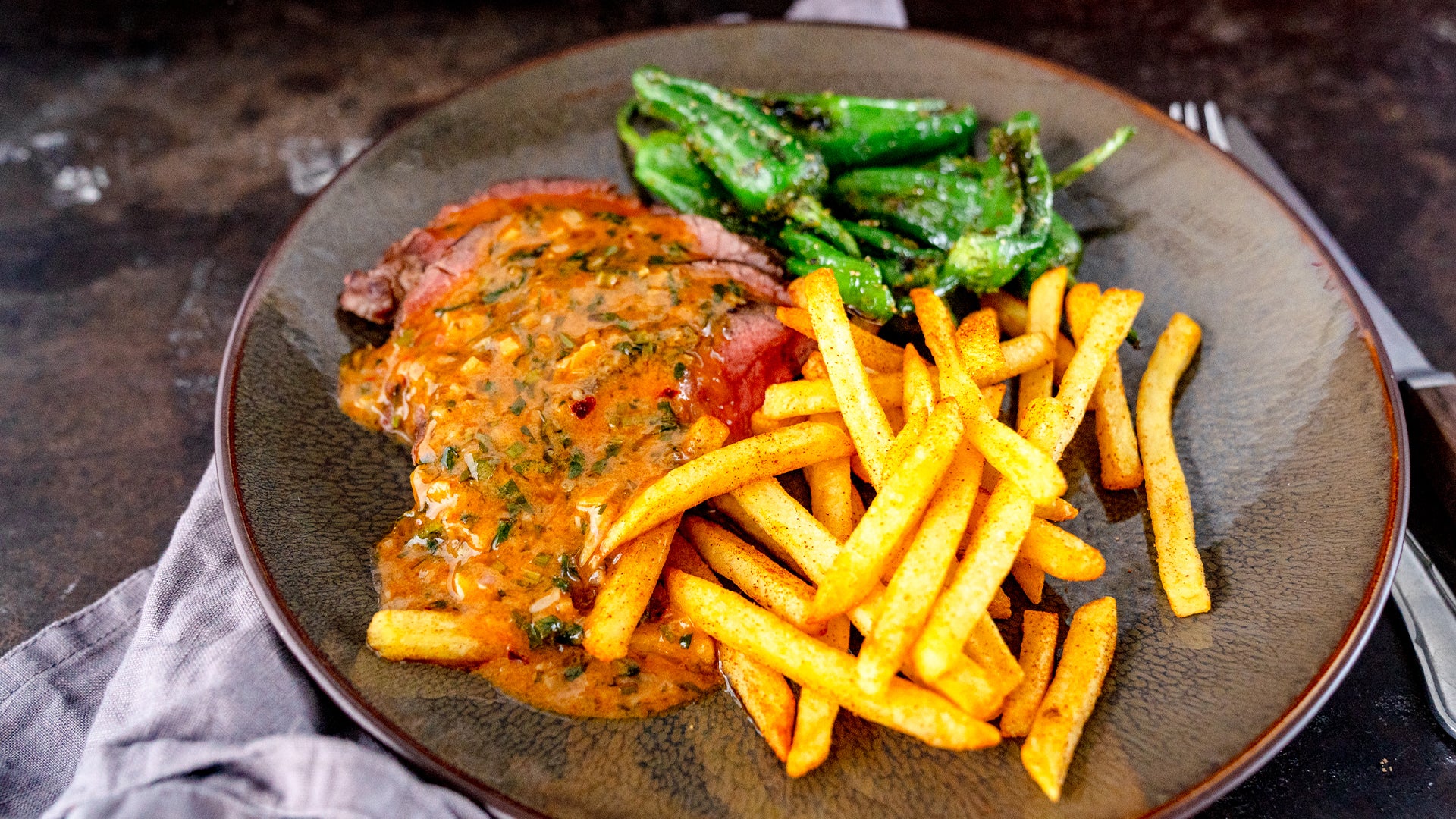 Ein Teller mit Steakscheiben, garniert mit Kräutersauce, goldbraunen Pommes Frites und gerösteten grünen Paprikaschoten, serviert auf einer dunklen Keramikschale, daneben ist teilweise eine graue Serviette zu sehen.