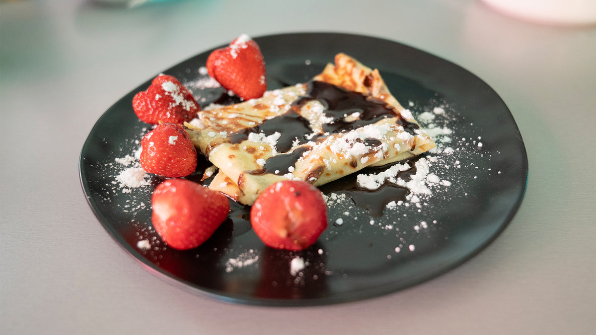 Ein schwarzer Teller mit gefalteten Crêpes, beträufelt mit Schokoladensauce, umgeben von frischen Erdbeeren und bestreut mit Puderzucker.