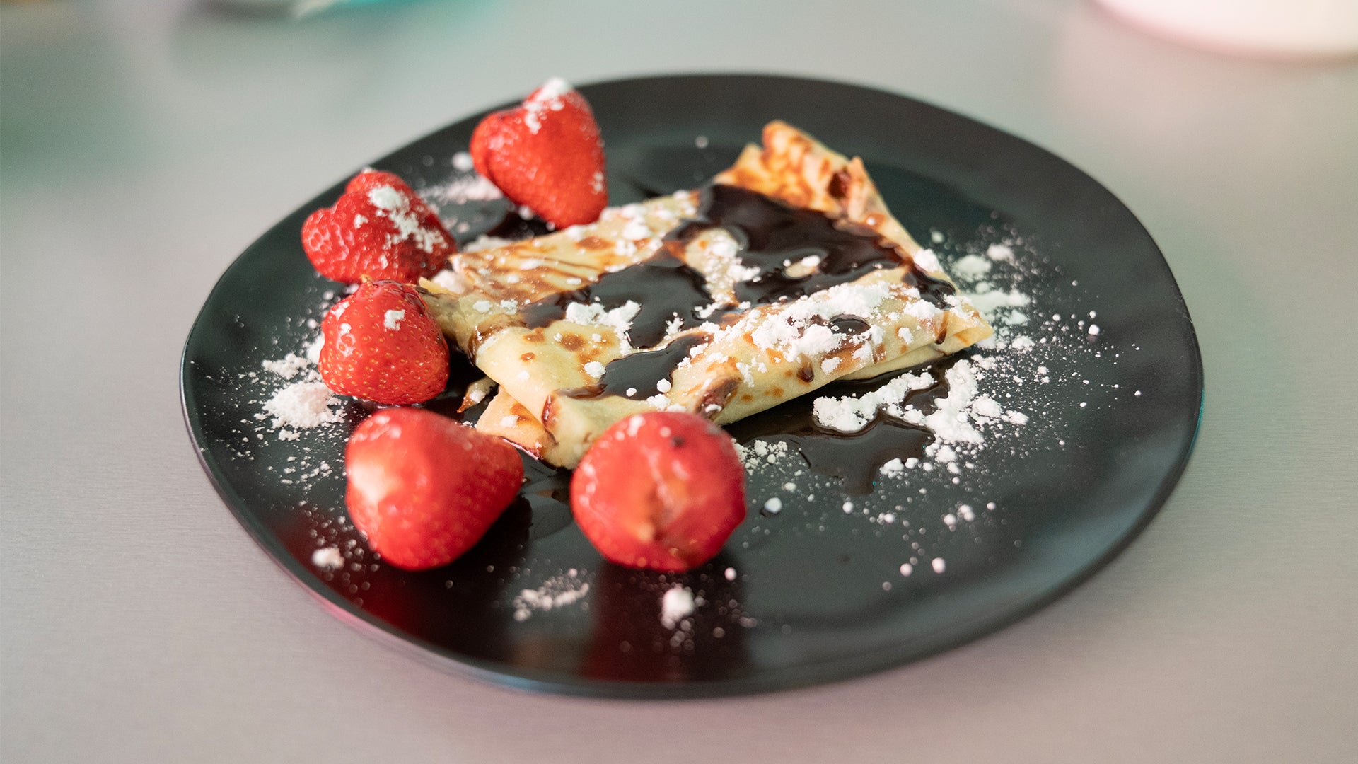 Ein schwarzer Teller mit gefalteten Crêpes, beträufelt mit Schokoladensauce, umgeben von frischen Erdbeeren und bestreut mit Puderzucker.