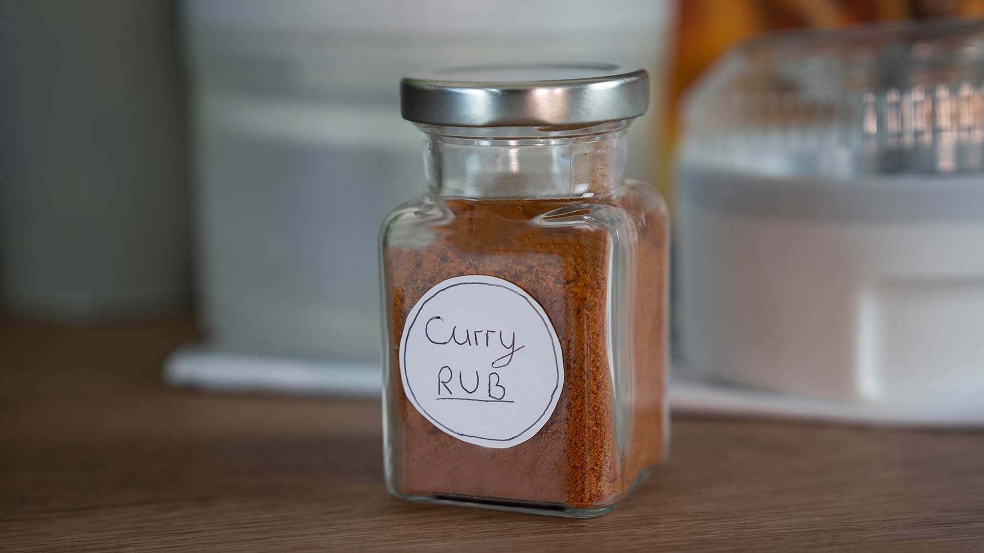 Ein Glasgefäß mit silbernem Deckel, gefüllt mit einer rotbraunen Gewürzmischung und der Aufschrift „Curry Rub“, steht auf einer Holzoberfläche, im Hintergrund sind unscharfe Küchenutensilien zu sehen.