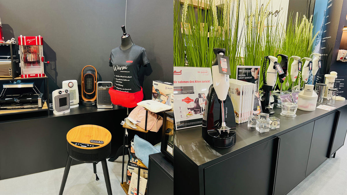 Eine Ausstellung von Haushaltsgeräten und Gadgets auf schwarzen Theken, darunter eine Schaufensterpuppe mit einer Markenschürze, ein Lautsprecher mit Holzplatte, Heizgeräte, Reinigungsgeräte und verschiedene Produktkartons, mit grünem Ziergras im Hintergrund.