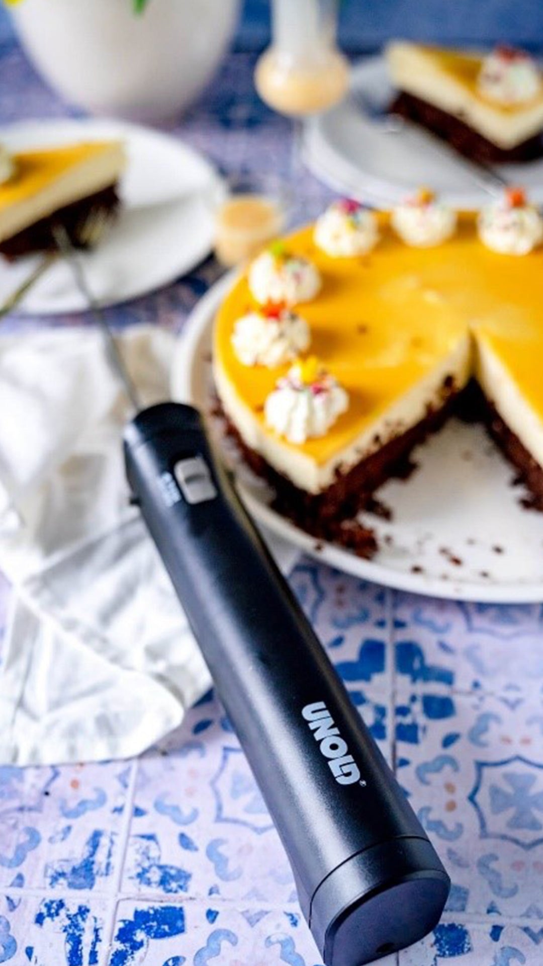 Ein schwarzes elektrisches Messer liegt auf einer blau gemusterten Oberfläche neben einem teilweise angeschnittenen Kuchen mit gelbem Belag und Schlagsahne-Dekoration, im Hintergrund liegt ein Kuchenstück auf einem Teller.