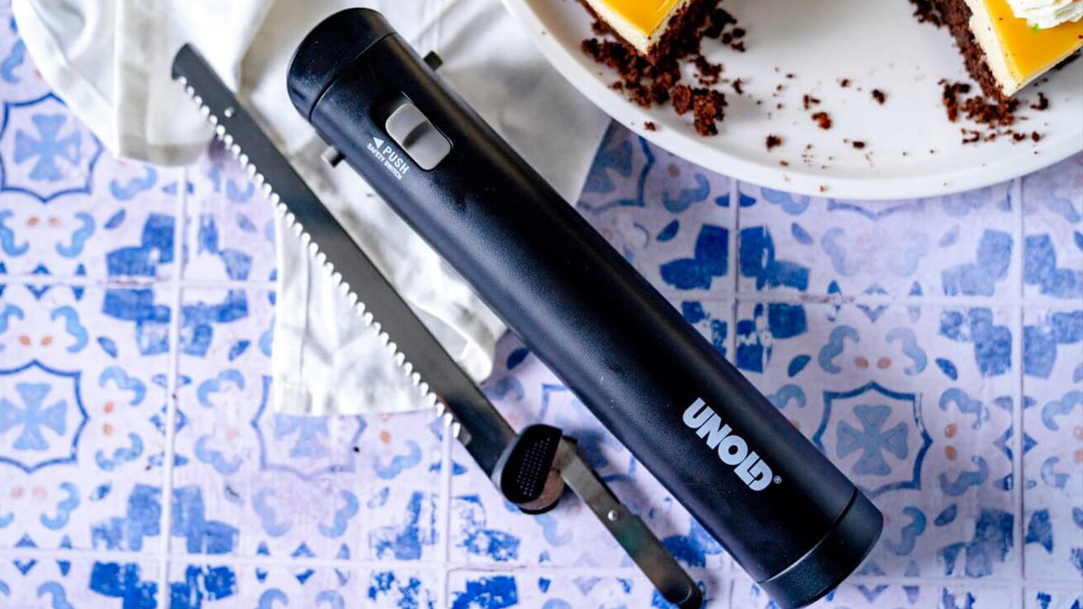 Ein schwarzes elektrisches Messer mit gezackter Klinge und der Aufschrift „UNOLD“ liegt auf einer blau gemusterten Oberfläche neben einer weißen Serviette und einem Teller mit teilweise geschnittenen Kuchenstücken.